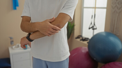 Obraz premium Young man clutching elbow in pain at gym with exercise equipment visible, demonstrating discomfort and exercise-related strain.