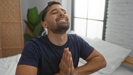 Young man smiling joyfully with hands clasped while sitting on a bed in a peaceful bedroom setting,...