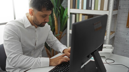 Fototapeta premium Young man in white shirt working at a computer in a modern office space with indoor plants and bookshelves, focusing on his task in a professional environment.