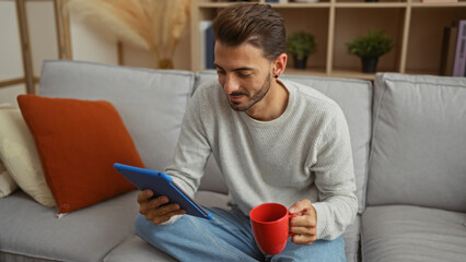 Young hispanic man sitting in living room using tablet with red mug wearing casual sweater and jeans relaxed on comfortable couch in cozy home interior