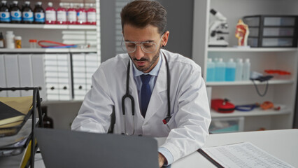 Handsome hispanic man working on laptop in a modern clinic's interior with medical equipment around, conveying a professional and focused atmosphere.