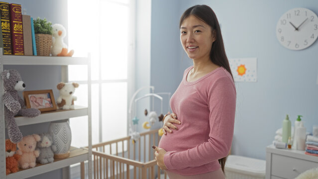 Pregnant woman in pink sweater stands smiling in cozy baby nursery with crib, soft toys, books, and natural light creating a warm, inviting atmosphere
