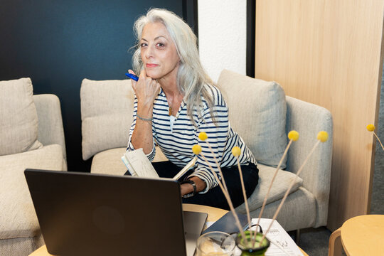 Senior Woman Thinking With Notebook And Laptop In Office
