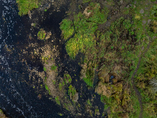 Top-down aerial photo of the Jagala River near Jägala Waterfall in Estonia. On the left the river flows with dark, almost black water speckled with white foam. On the right rugged green landscape.