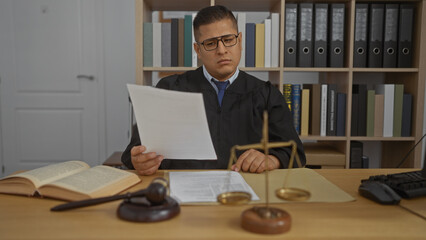 Male judge working in an office with legal books and scales of justice on the desk while reading...