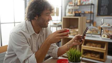 Young man enjoying coffee and using his smartphone in a cozy bakery with a variety of baked goods in the background.