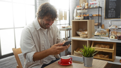 Young man smiling while looking at his phone in a cozy bakery, surrounded by delicious pastries and a coffee on the table.