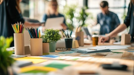 Office desk filled with colored pencils, plants, notepad, and sticky notes, while creative professionals collaborate on projects in the blurred background