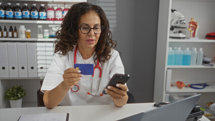 Woman in clinic holding credit card and smartphone wears glasses and stethoscope surrounded by medical supplies and white uniform focused on online transaction work