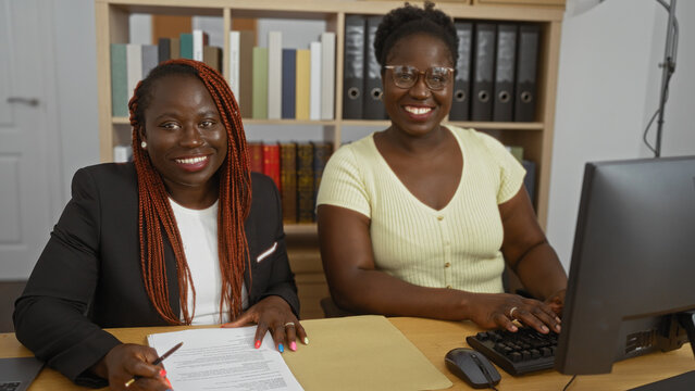 African american women business partners working in an office, one typing on a computer and the other taking notes.