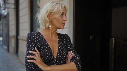 Fototapeta premium Beautiful blonde woman with short hair stands with arms crossed on an urban street, looking serious and pensive in a stylish city environment.