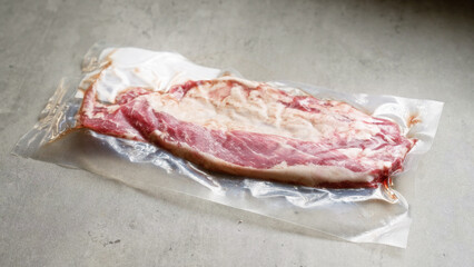 Vacuum-sealed raw bacon slice laying on a gray concrete surface showcasing packaging and freshness.