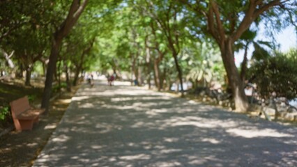 Blurred outdoor pathway with defocused trees and people walking in the background under sunlight creating bokeh effects in a park setting with shadows and greenery around