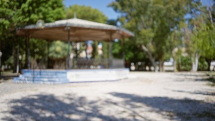 Outdoor park scene with defocused gazebo surrounded by green trees and sunny sky, blurred background highlighting bokeh effect and natural light