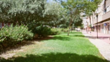 Obraz premium Blurred image of an outdoor park with trees, grass, and buildings in the background on a sunny day.
