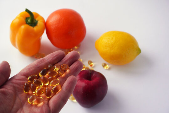 A hand holding several shiny golden capsules is extended in front of a vibrant arrangement of a yellow bell pepper, an orange, a lemon, and a red apple on a white background.