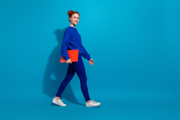 Candid portrait of a red-haired young woman in blue attire walking confidently with a red folder against a blue background