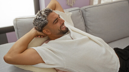 Handsome young arab man with a beard relaxing on a sofa at home, wearing a white shirt and smiling in a modern living room.