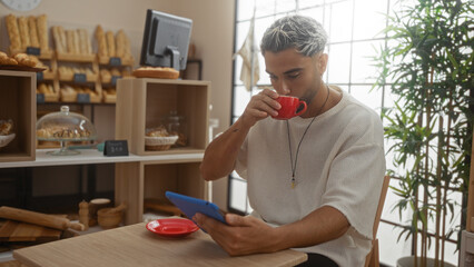 Young man with beard sits in a cafe drinking coffee and using a tablet, surrounded by bread and pastries in a cozy bakery setting.