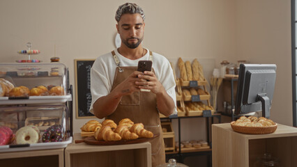Young man with a beard using a smartphone in a bakery shop surrounded by pastries and bread displays.