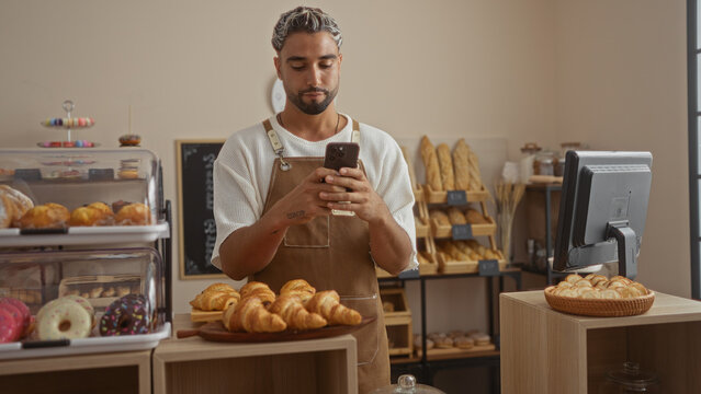 Young man with a beard using a smartphone in a bakery shop surrounded by pastries and bread displays. - Powered by Adobe