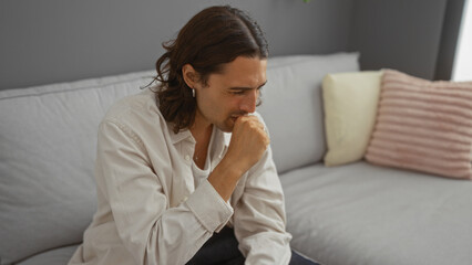 Young man coughing indoors while sitting on a gray couch in a modern living room