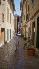 Woman walking through a charming alley in fornalutx, mallorca, spain, wearing white clothes and holding a basket on a sunny day, surrounded by traditional buildings and cobblestone streets.