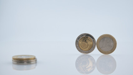 A 5 Zloty coin and a 1 Euro coin displayed on a reflective white surface with additional stacked coins. 
