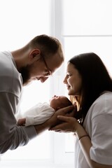 Portrait of a young family with infant in his arms photographed on a light window background.