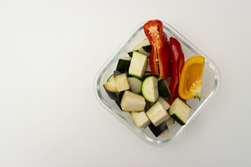 Glass container with chopped vegetables on white background. Zucchini, eggplant and bell pepper for baking. Ingredients for dinner. Top view, copy space