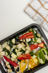 On a white table there is a metal baking tray with vegetables. Close-up, copy space. Zucchini, bell pepper and eggplants for baking.