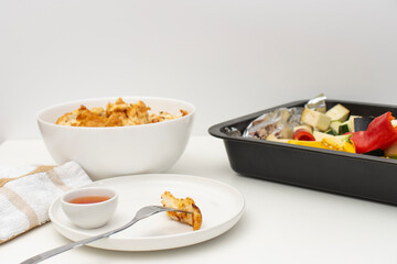 On a white table there is a bowl with fried chicken, a baking tray with vegetables and a plate with a fork. Homemade dinner on a white background. Copy space