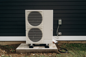 A large white outdoor air conditioner stands on a betaine base against a black wooden wall until it is hung in the air conditioner space.