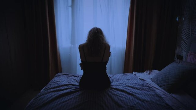 black silhouette upset woman sitting on bed dark room, suffer from insomnia, difficulty falling asleep, sleep problems. Sad lonely Girl feels bad. window curtains white fabric fly in wind blowing 4k.