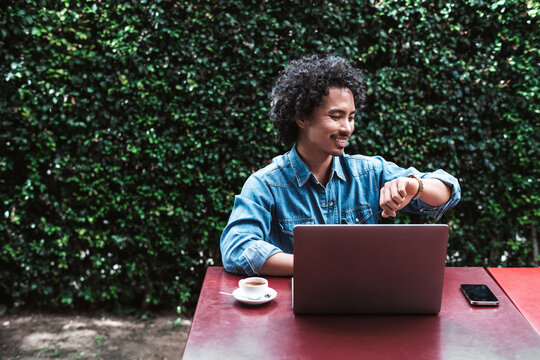 Smiling young man with curly hair sitting outdoors at a red table, working on a laptop, checking his watch, with a smartphone and coffee cup nearby, green hedge background - Powered by Adobe