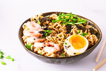 Ramen noodles with egg, shrimps, greens, seasame, seaweed and spices on white marble table close up. Traditional Asian soup.
