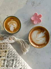 Two lattes with delicate foam art bask in the soft spring sunlight in Istanbul, accompanied by a crocheted decoration and a pink flower clip on a marble table