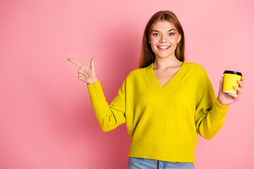 Young woman in yellow sweater holding coffee cup and pointing to empty space on pink background showcasing lifestyle and positivity