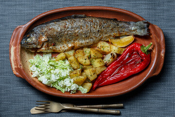 Tasty baked trout fish with red pepper, potatoes and Chinese cabbage salad in a clay dish on the table, closeup, top view
