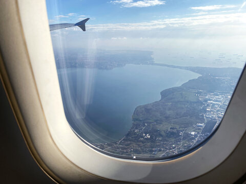 A picturesque aerial view of a coastline and water body captured through an airplane window, featuring the aircraft wingtip and a vibrant blue sky with scattered clouds. - Powered by Adobe