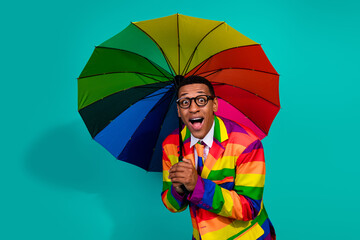 Young man in a rainbow-striped suit with a matching colorful umbrella on a teal background.