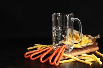 Empty glass for beer or kvass and snacks on dark background. Traditional glass beer mug with handle. Close-up of glass on wooden board and French fries, hunting sausages