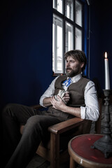 A pensive man in vintage attire clutches an old book, seated in a dimly lit room with blue walls and a single candle, evoking a somber and historical atmosphere.