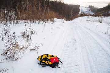 A bright yellow and black backpack is carelessly laying on a snowy road, surrounded by a...