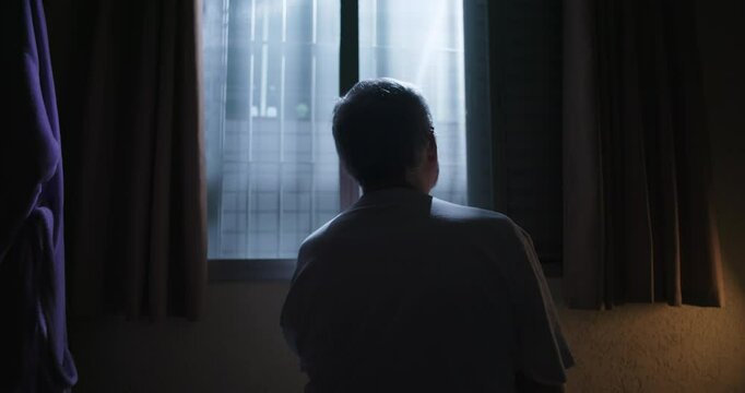 Elderly man sitting in a dark room, facing a window illuminated by moonlight, enveloped in quiet solitude and profound introspection