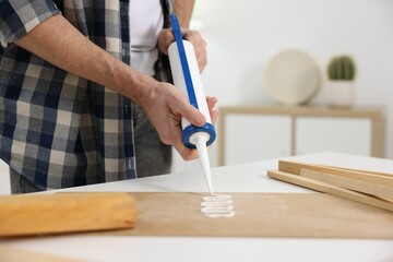 Man with caulking gun glueing plywood at white table indoors, closeup