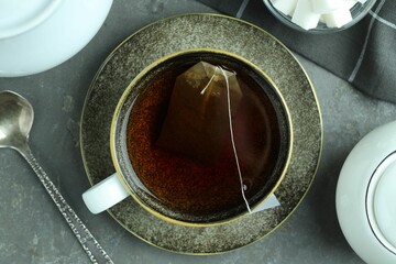Tea bag in cup with hot drink, teapot, sugar cubes and spoon on gray textured table, flat lay
