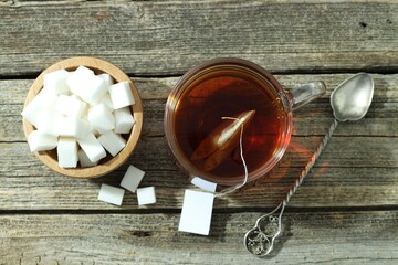 Tea bag in glass cup with hot drink, sugar cubes and spoon on wooden table, flat lay