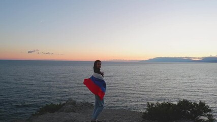 A brunette woman with a Russian flag on her shoulders stands on a rocky seashore at sunset. Gives independence, patriotic holiday concept - Powered by Adobe