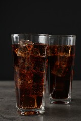 Cold cola with ice cubes in glasses on grey table against black background, closeup